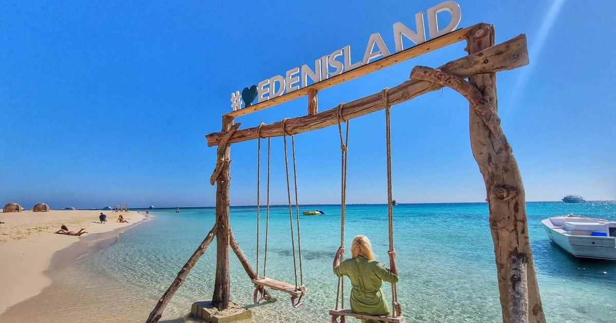 Drewniana huśtawka pod napisem EDEN ISLAND na białej piaszczystej plaży Wyspy Eden, kobieta patrzy na turkusowe Morze Czerwone z zacumowanymi łodziami