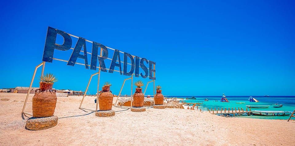 Napis PARADISE na białej plaży Wyspy Paradise z turkusowym morzem i łodziami w tle