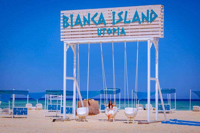 Biała huśtawka z napisem Bianca Island Utopia na piaszczystej plaży z niebieskimi parasolami nad turkusowym Morzem Czerwonym
