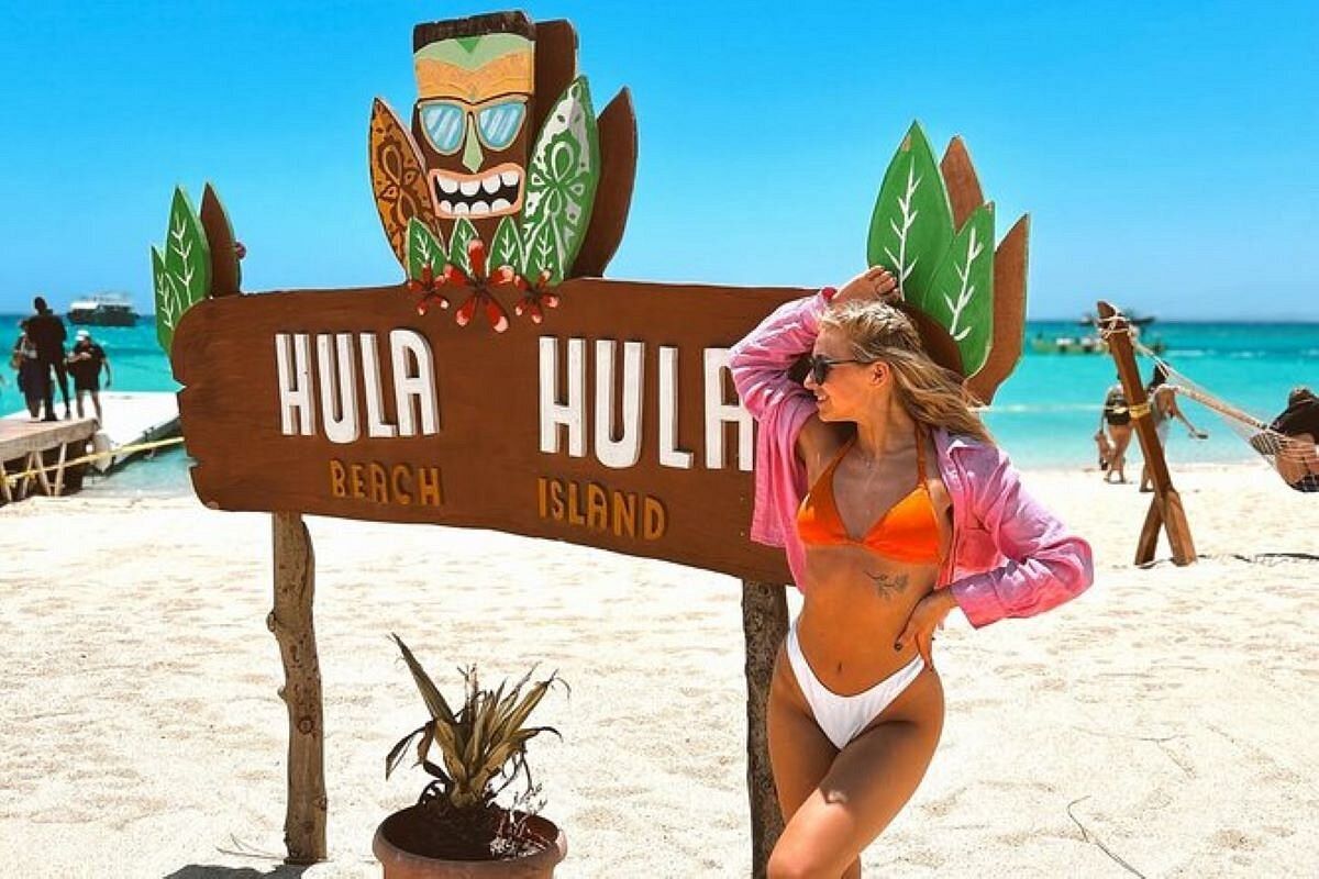 Drewniana tablica Hula Hula Beach Island na białej plaży w Hurghadzie z turkusowym Morzem Czerwonym i łodziami w tle
