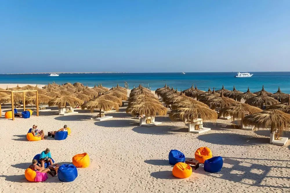 Plaża z rzędami słomianych parasoli oraz pomarańczowymi i niebieskimi pufami na białym piasku nad turkusowym morzem koło Hurghady