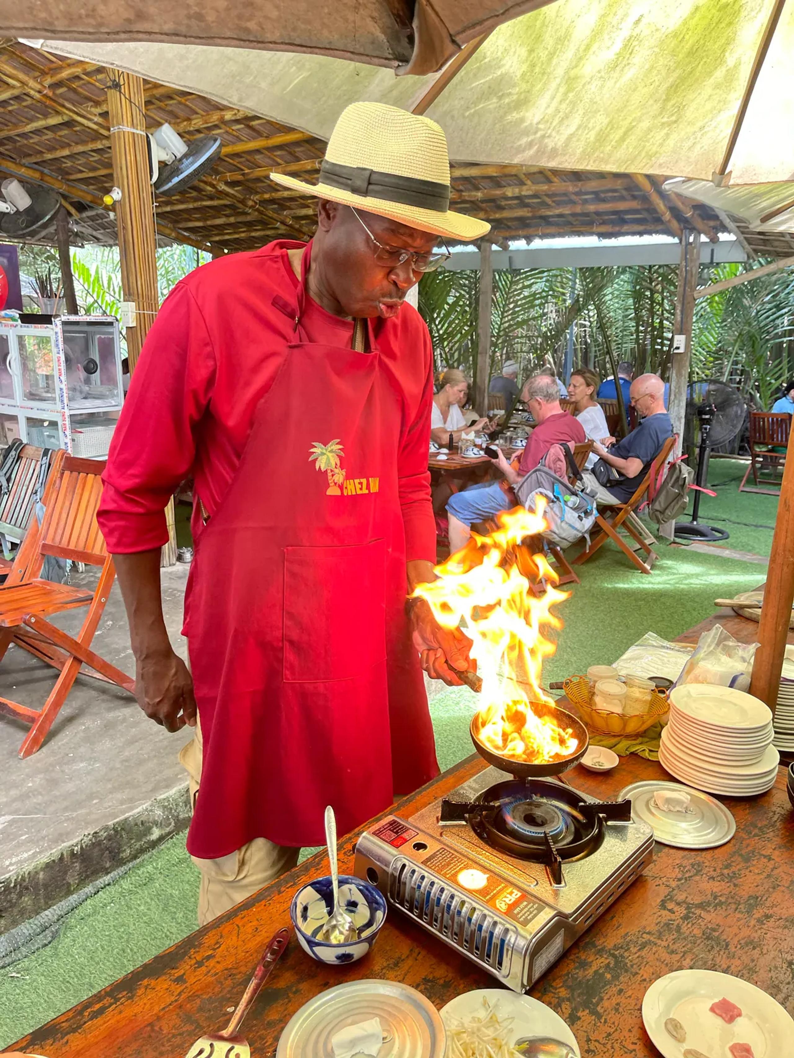 A guest cooking over an open flame in an apron and chef's hat at the village kitchen