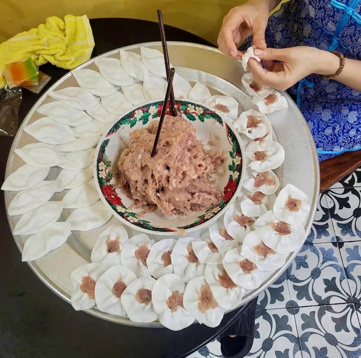Hands pinching white rose dumplings around a bowl of shrimp filling