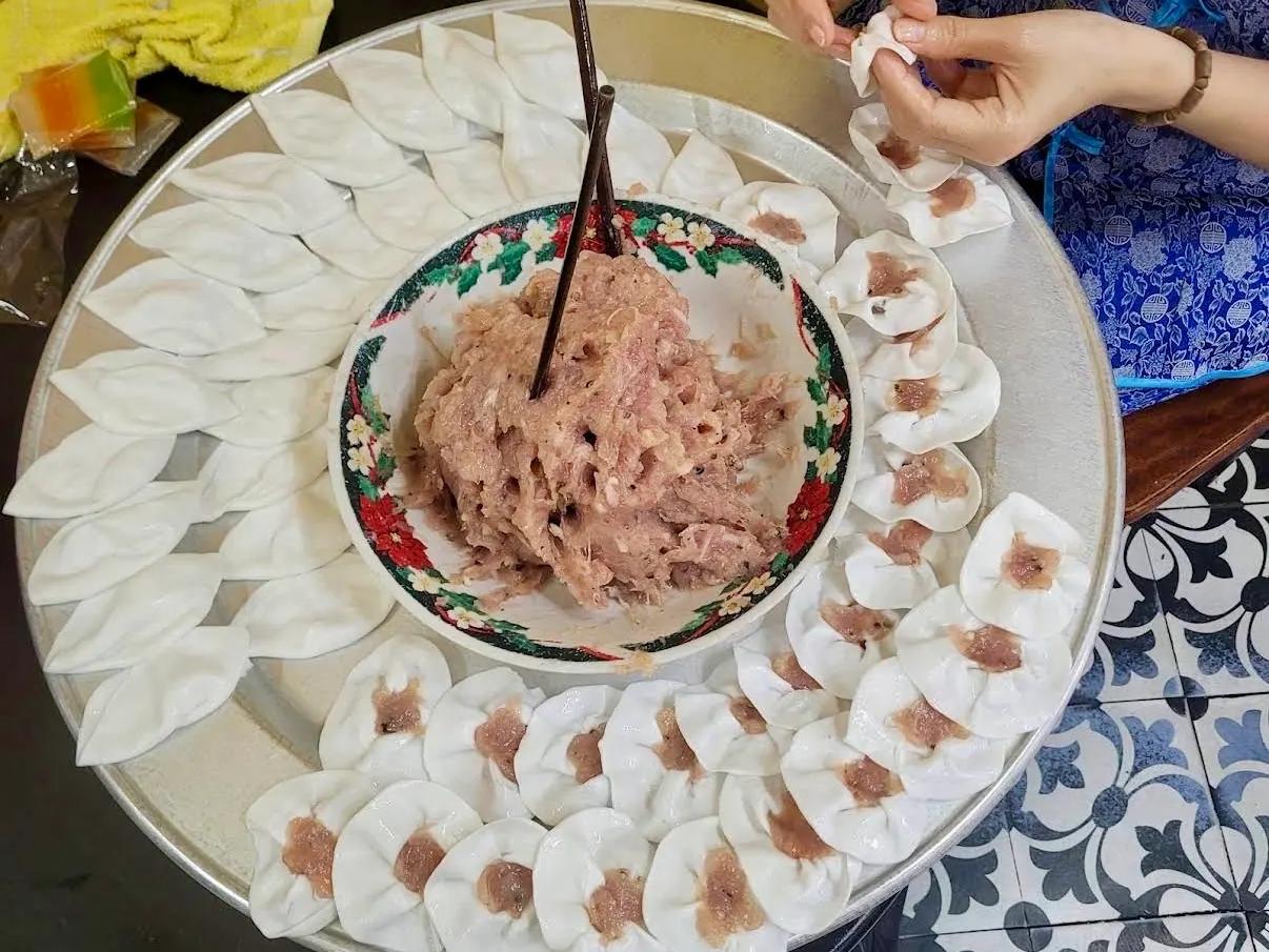 Hands pinching white rose dumplings around a bowl of shrimp filling
