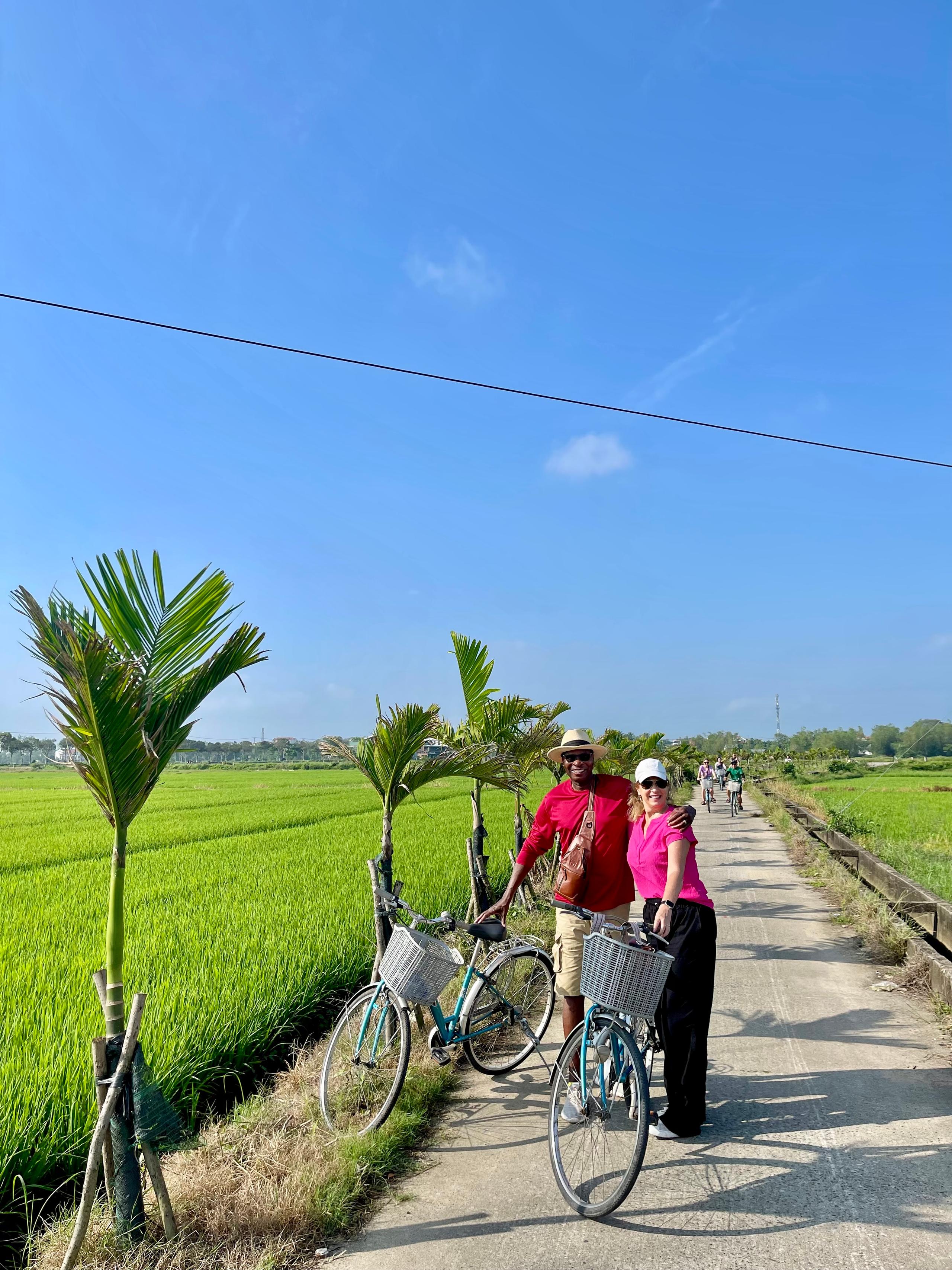 Two guests with their bicycles on a narrow path between green rice fields outside Hoi An