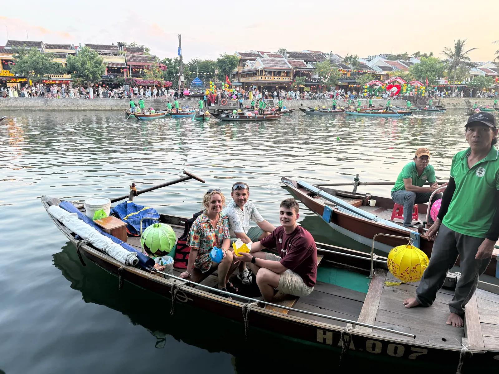 Three guests with paper lanterns in a wooden boat on the Hoai River at dusk, the Ancient Town riverfront behind