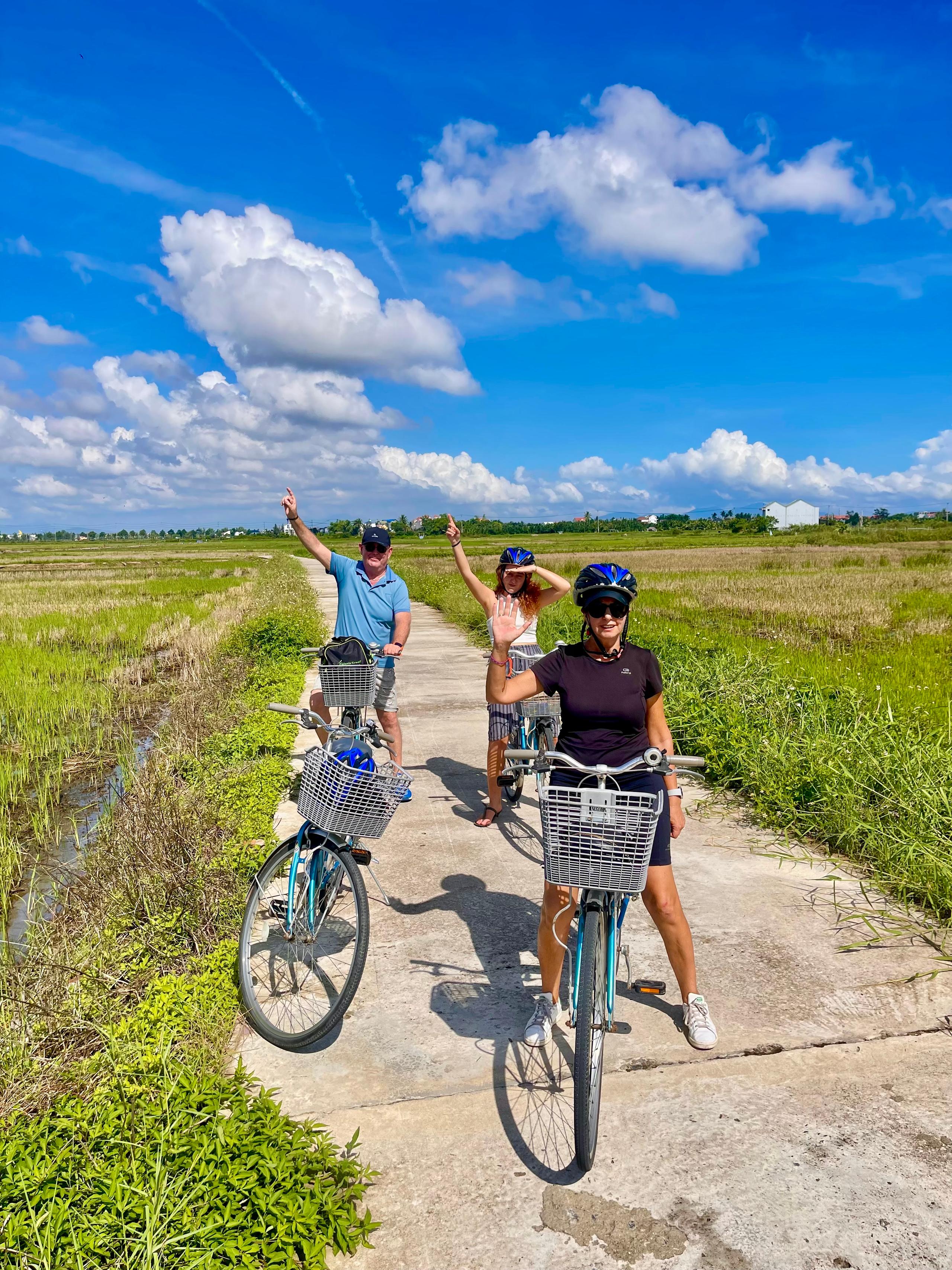 Three guests on bicycles waving from a narrow path through the rice fields outside Hoi An