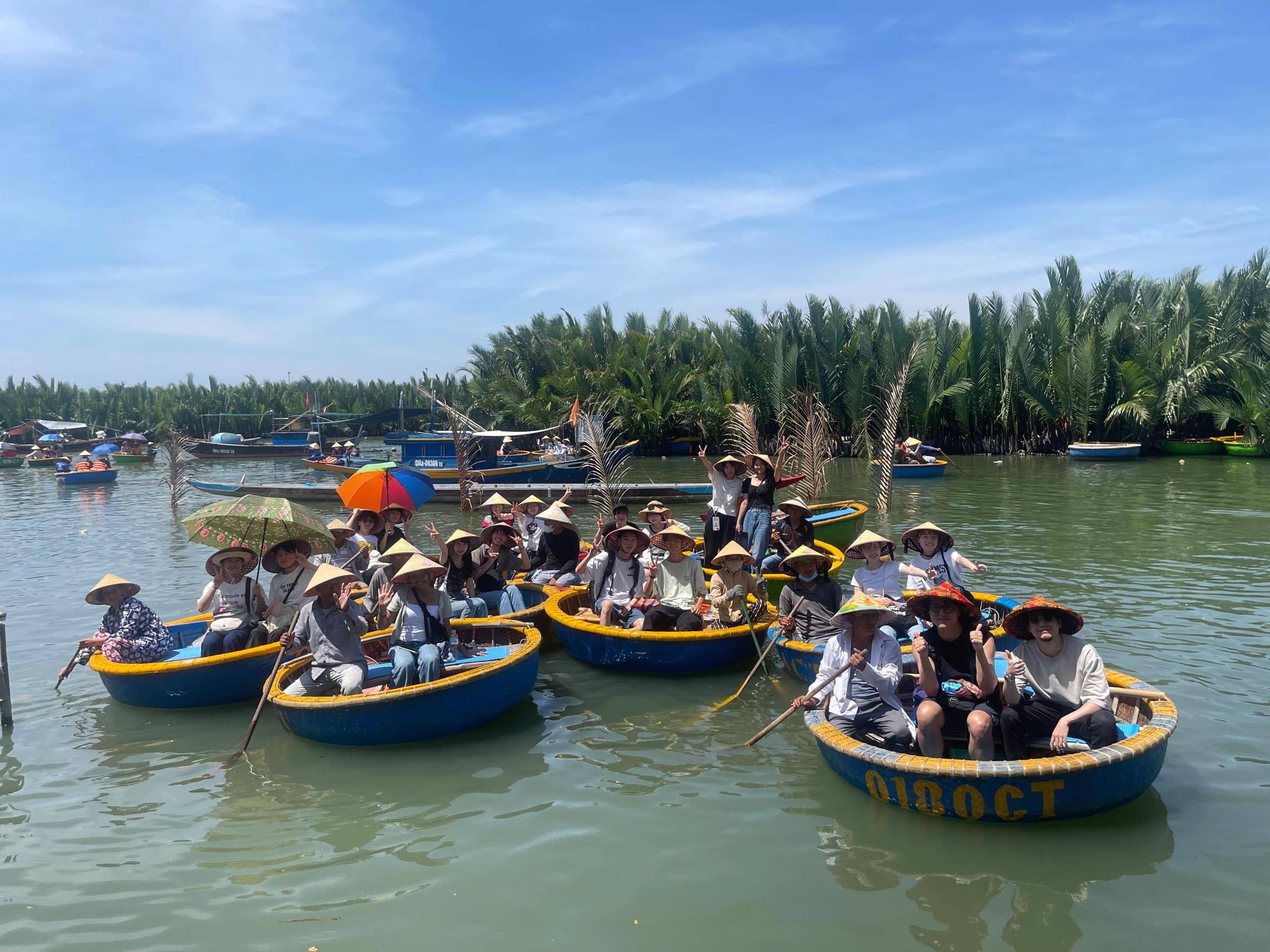 A large group in conical hats paddling round basket boats through the coconut palm forest near Hoi An