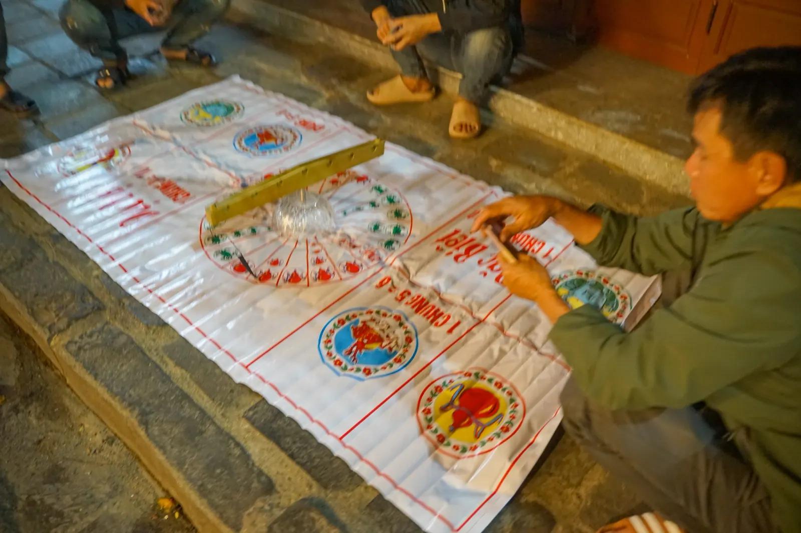A Bau Cua mat spread on the pavement at night, the glass bowl in the middle and players crouched around it