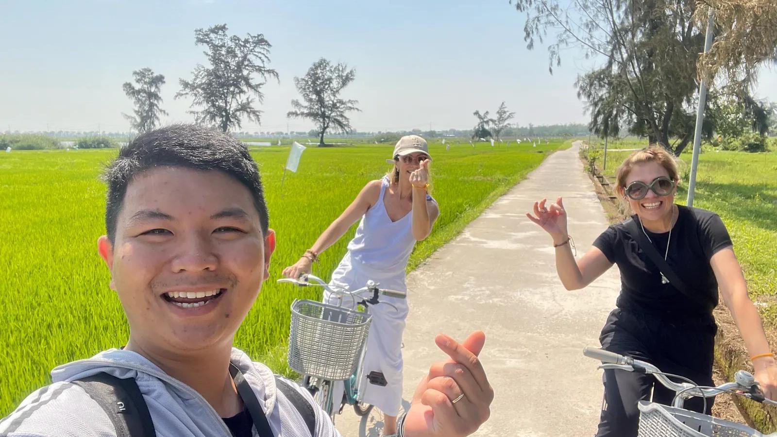 Tom cycling with guests along a concrete path between bright green rice fields outside Hoi An