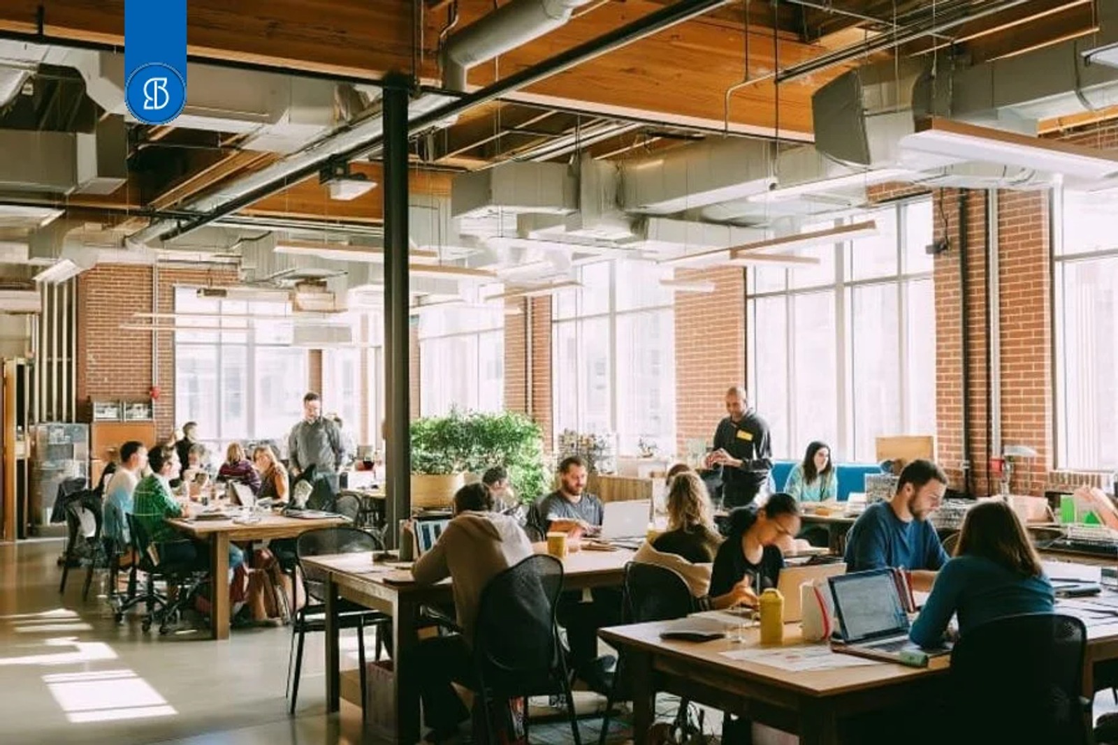 Espaço de coworking de tijolo aparente e teto industrial, com dezenas de pessoas trabalhando em mesas comunitárias