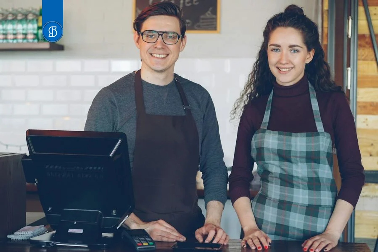 Dois donos de cafeteria sorrindo atrás do balcão, com terminal de ponto de venda e maquininha de cartão