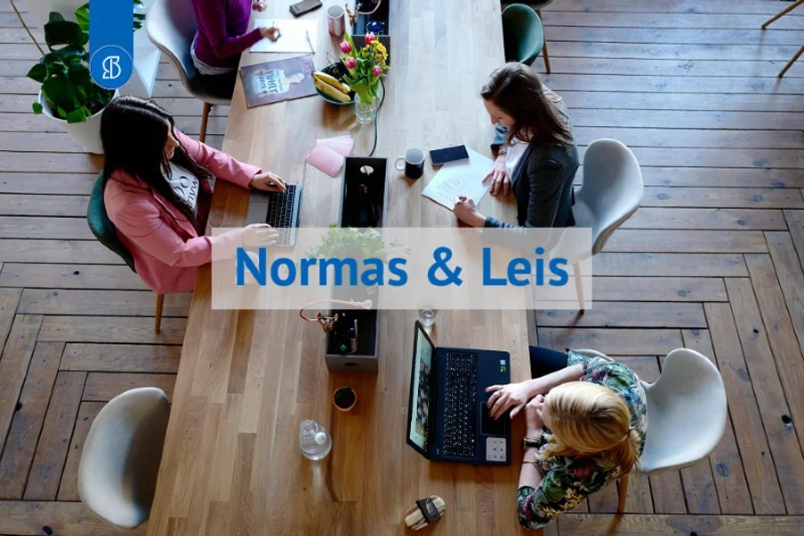 Vista de cima de três pessoas com notebooks em mesa de madeira, com a inscrição Normas e Leis