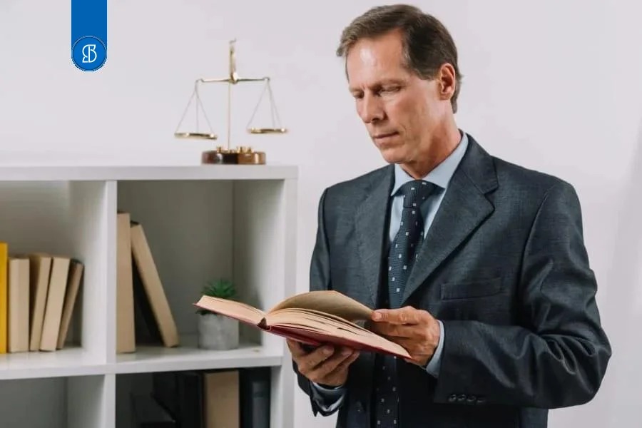 Advogado de terno cinza lendo um livro em pé, diante de estante com a balança da justiça