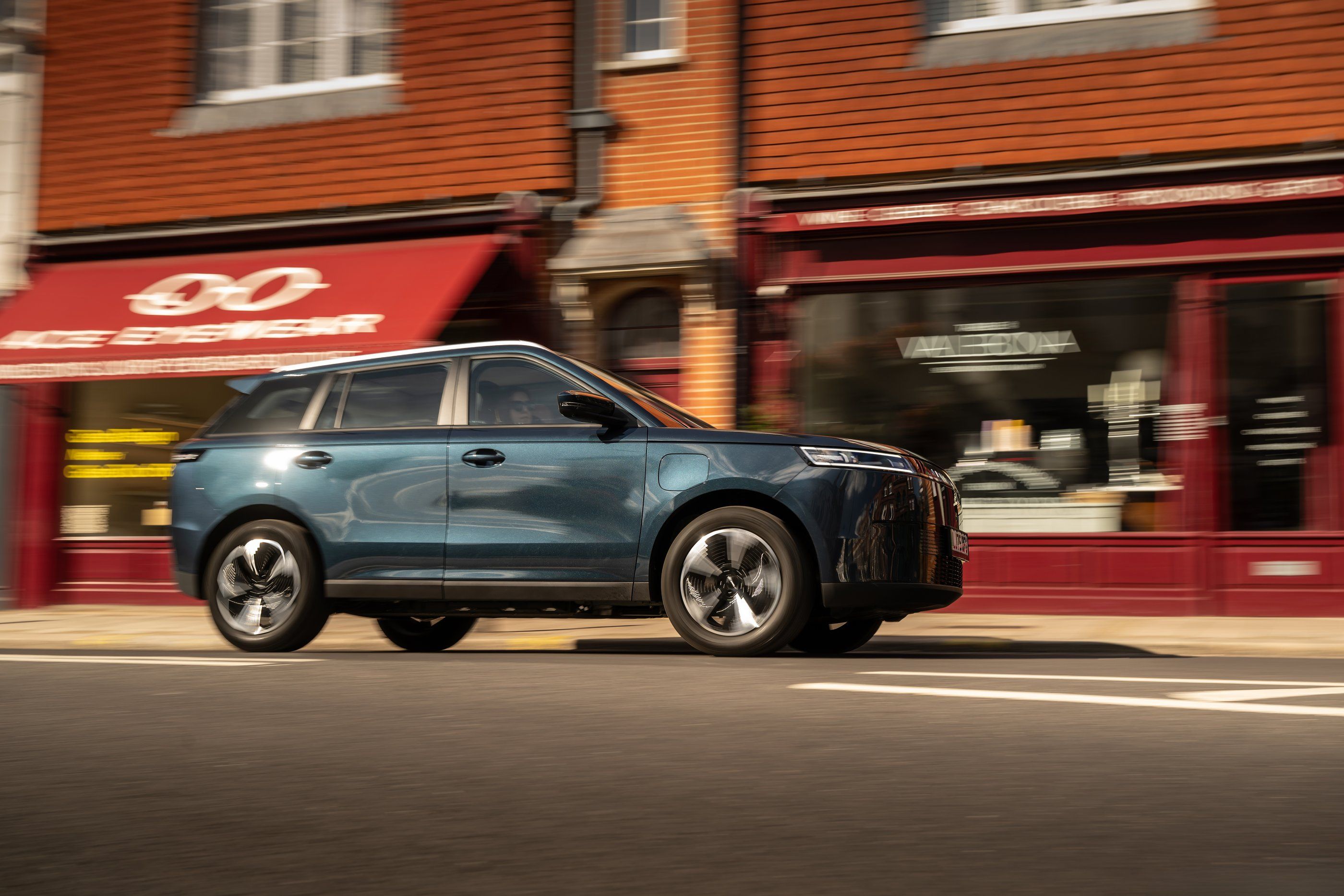 A blue JAECOO E5 driving past shops on a high street