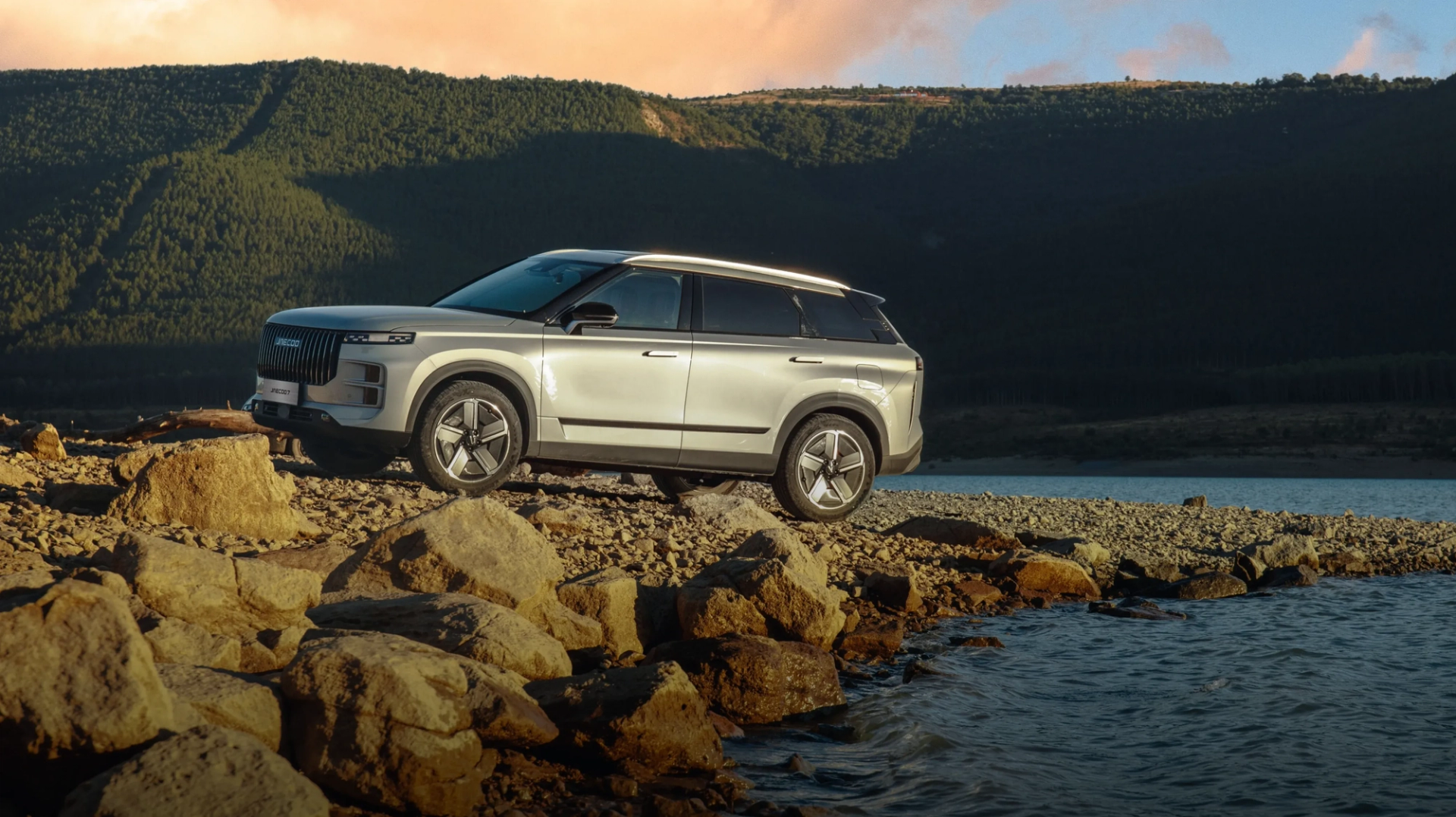 A silver JAECOO SUV parked on a rocky lakeside shore at dusk
