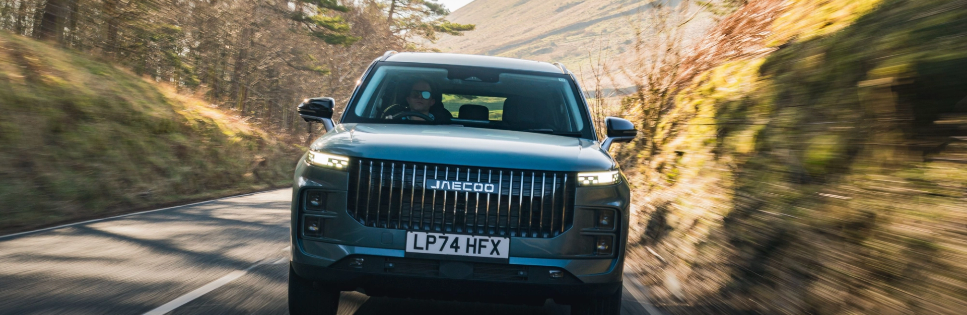 A JAECOO 7 driving head-on along a sunlit UK country road between wooded hillsides