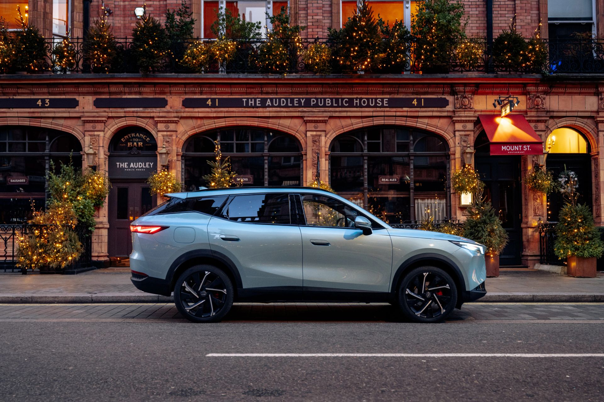 A silver OMODA SUV parked on a London street outside The Audley public house, lit with festive lights