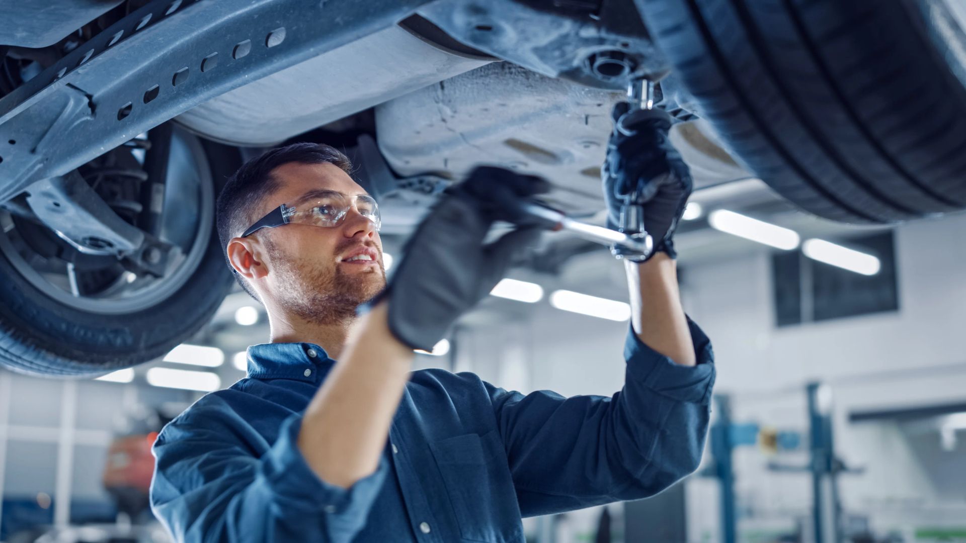 Mechanic in safety glasses tightens a bolt underneath a car on a workshop ramp
