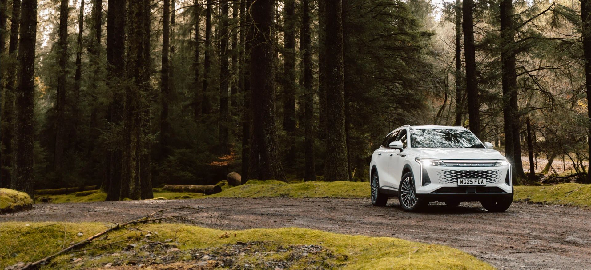 A white OMODA SUV parked on a forest track surrounded by tall pine trees