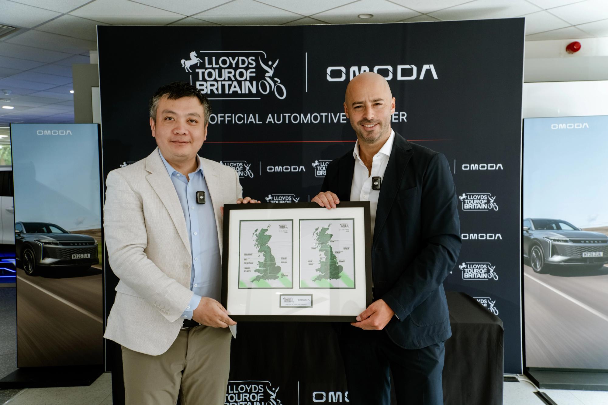 Image of two people holding up a map of Britain with Lloyds Tour of Britain and Omoda partnership board behind them in the background