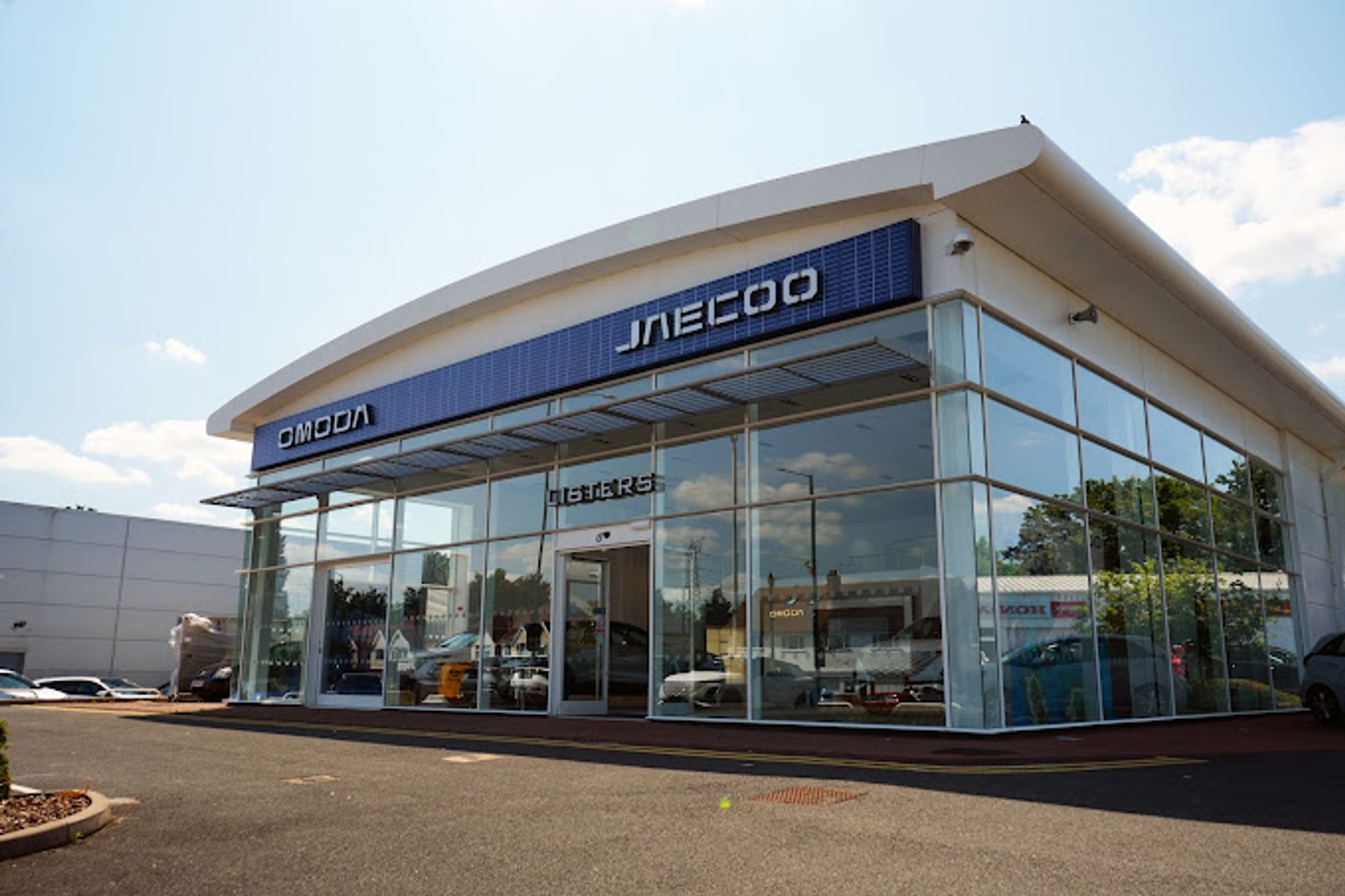 Glass-fronted OMODA and JAECOO dealership showroom with a curved roof and parked cars