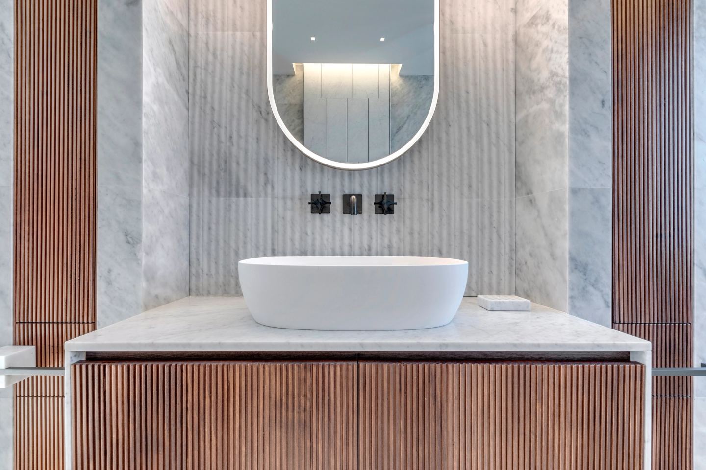 Fluted walnut vanity beneath the backlit oval mirror