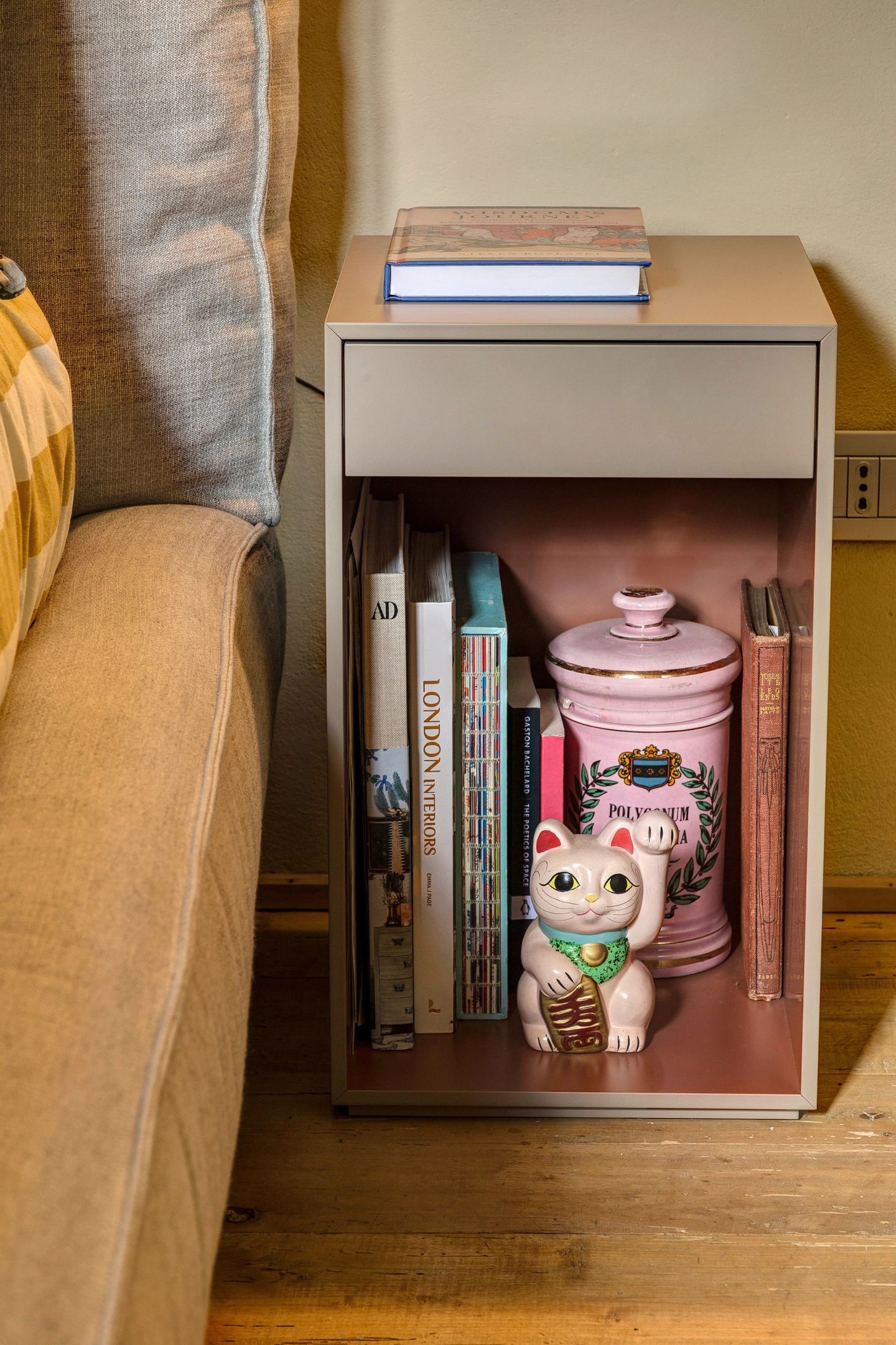 The bedside table's open shelf, pink inside, books and a lucky cat