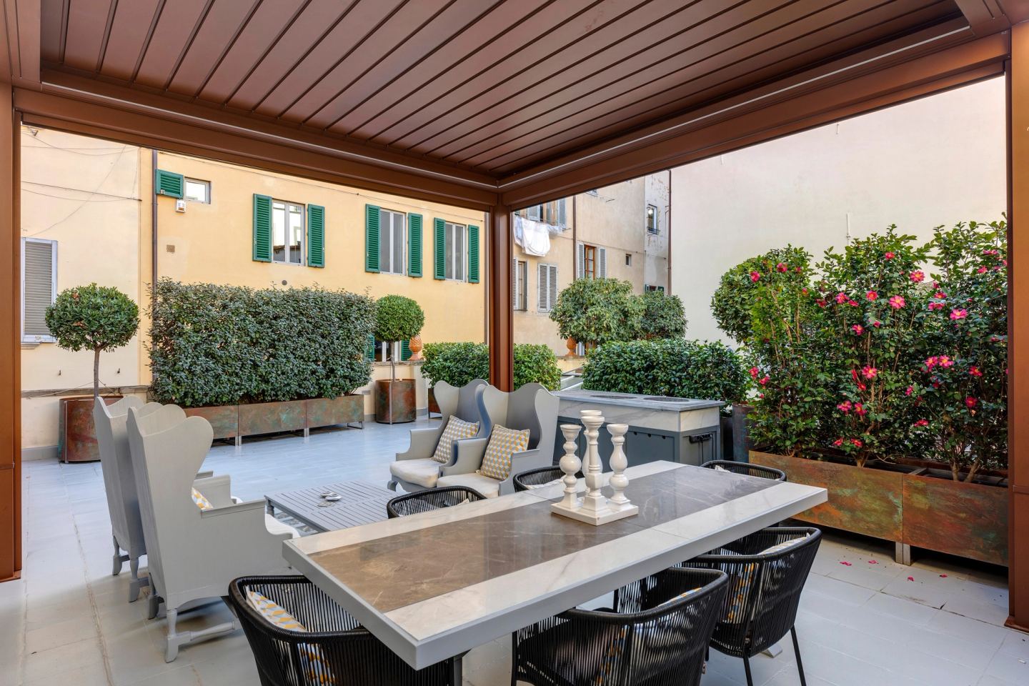 Il tavolo da pranzo sotto il tetto chiuso, sedie intrecciate intorno a un piano in marmo, siepi e arbusti fioriti alle spalle