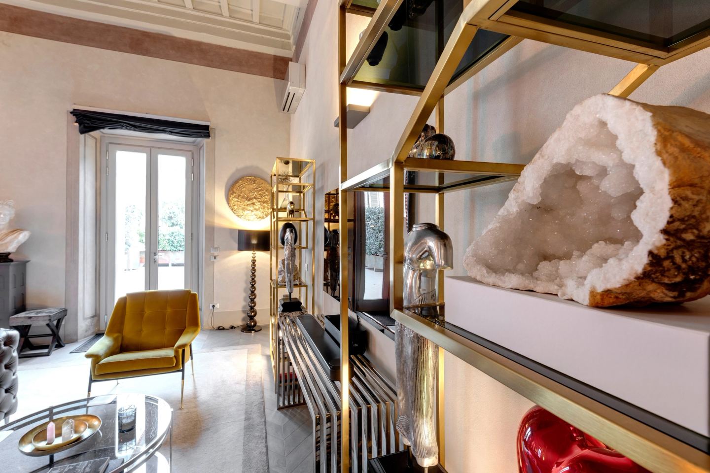 A gold shelving unit with a geode, a yellow armchair and a floor lamp by the window