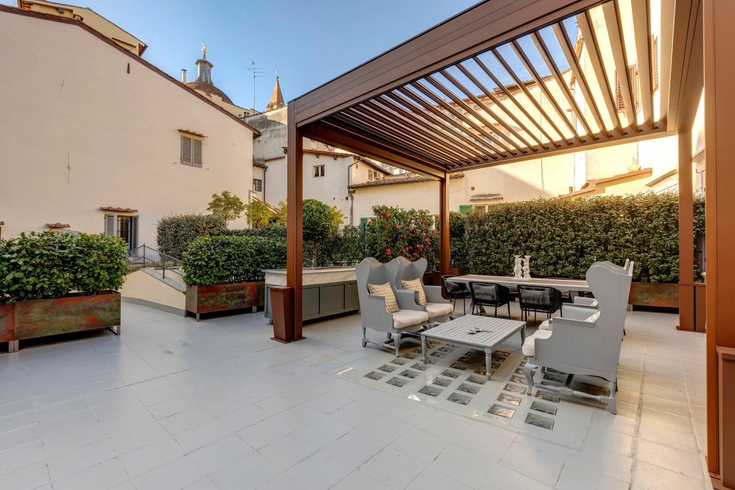 La terrazza nel sole del pomeriggio: la pergola in corten con le lame del tetto aperte, poltrone grigie a orecchioni e un tavolo da pranzo sotto