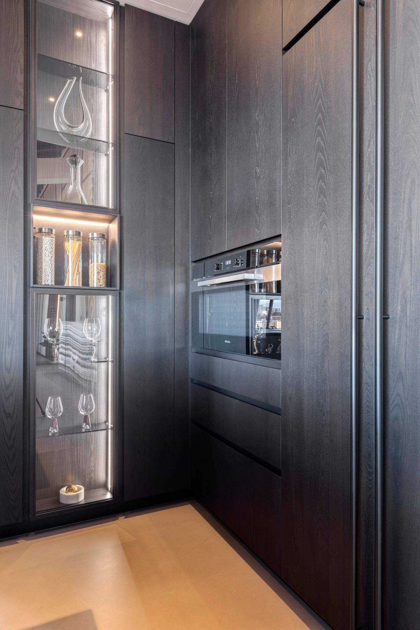 Tall dark cabinetry: oven, glassware, lit shelves