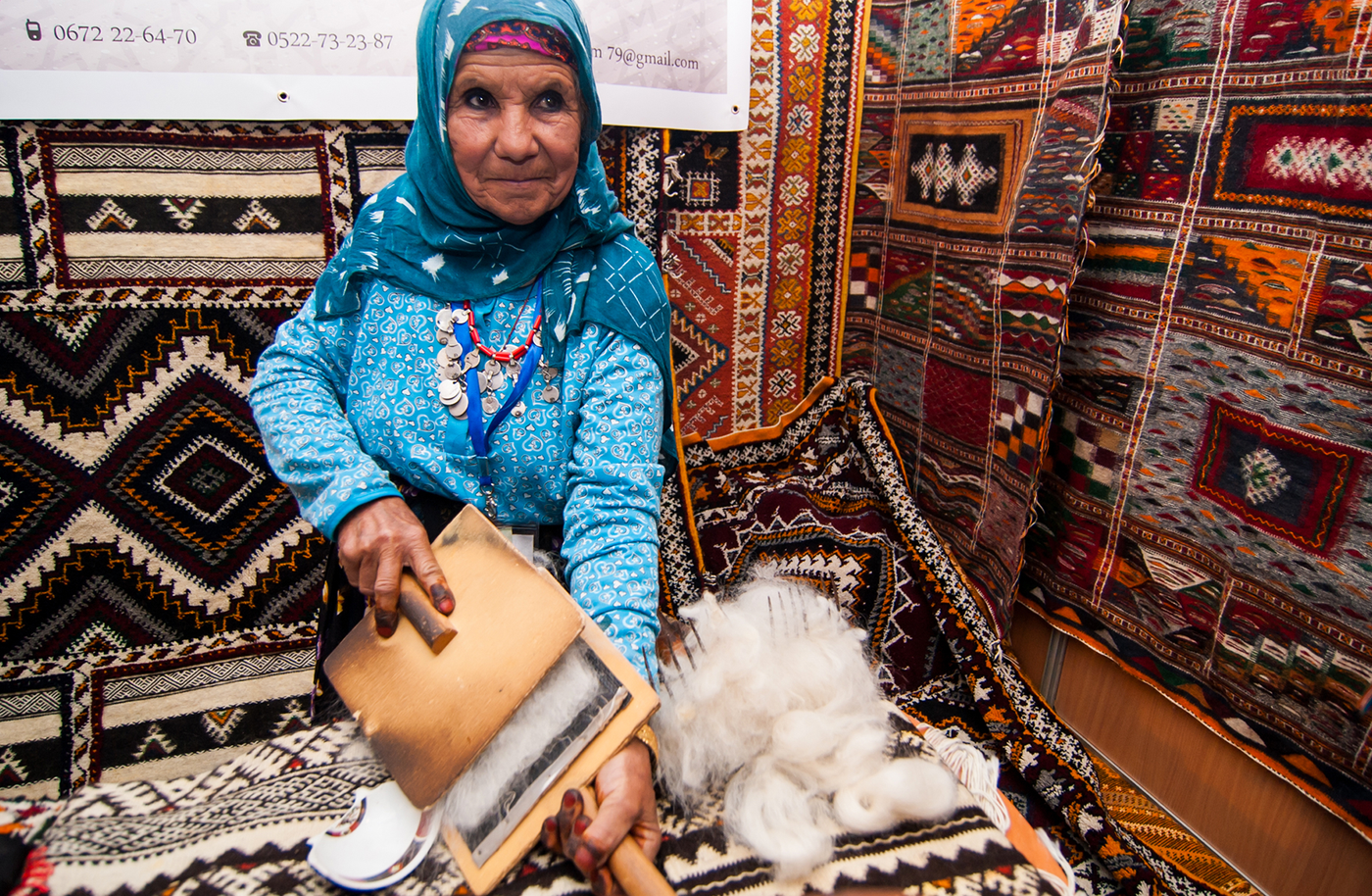 Elderly woman in blue carding raw white wool by hand, seated against stacked Berber carpets