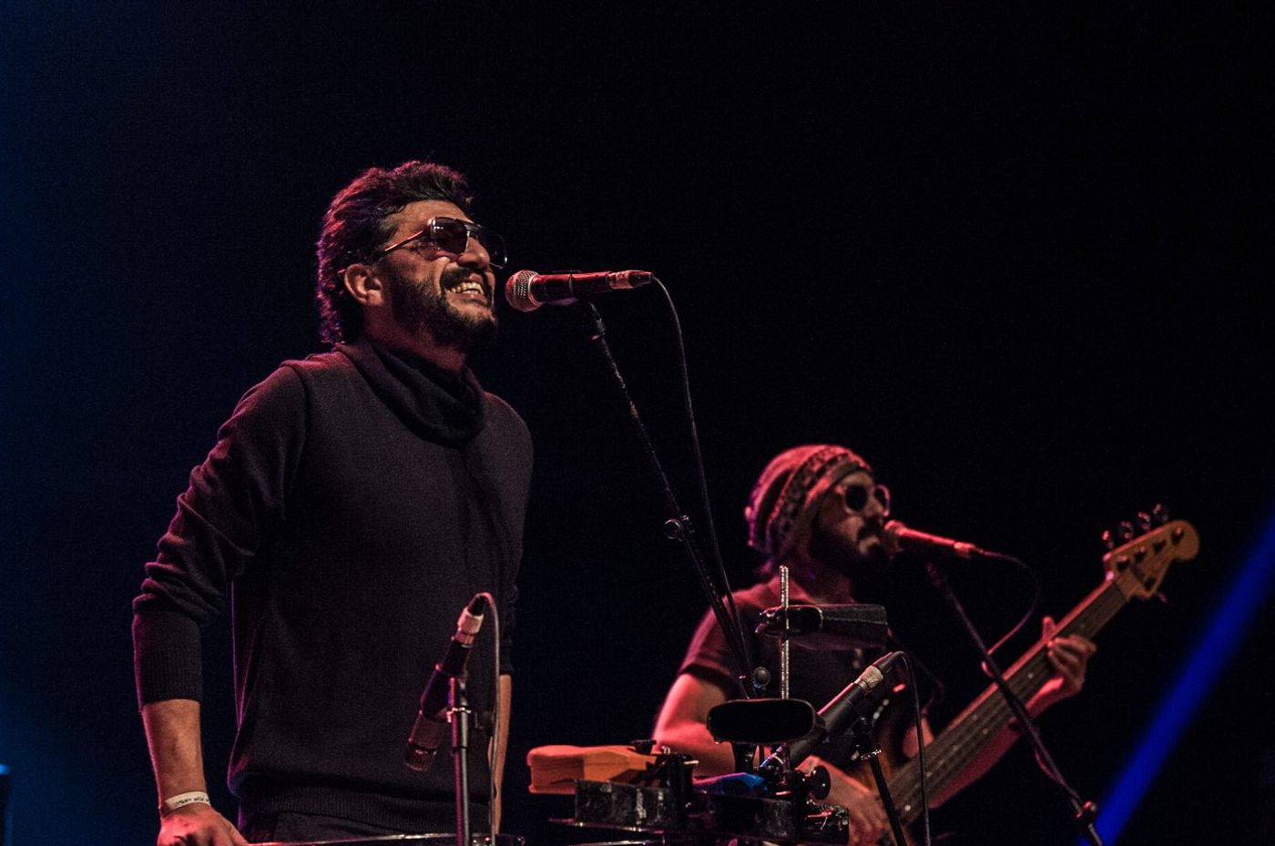 Bearded singer in sunglasses performs at a mic on a dark stage, bassist beside him