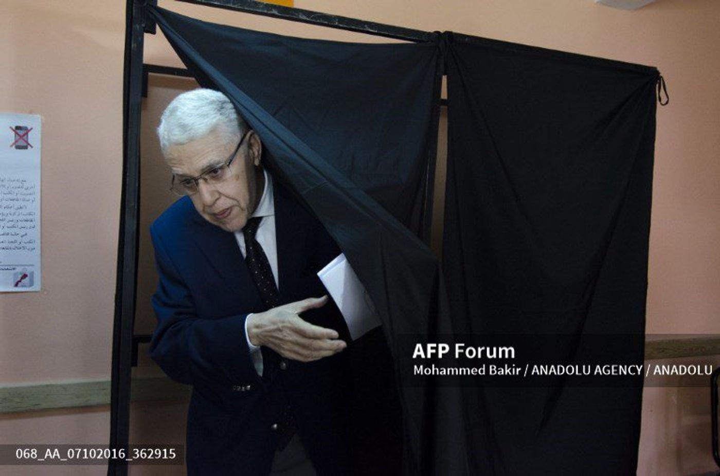 White-haired man in suit steps from a black voting booth curtain holding a ballot