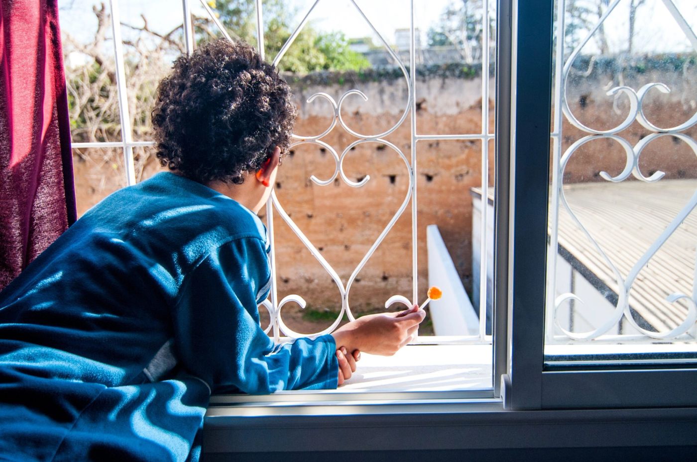 Curly-haired child in blue leans on a windowsill holding a lollipop, gazing through a wrought-iron grille
