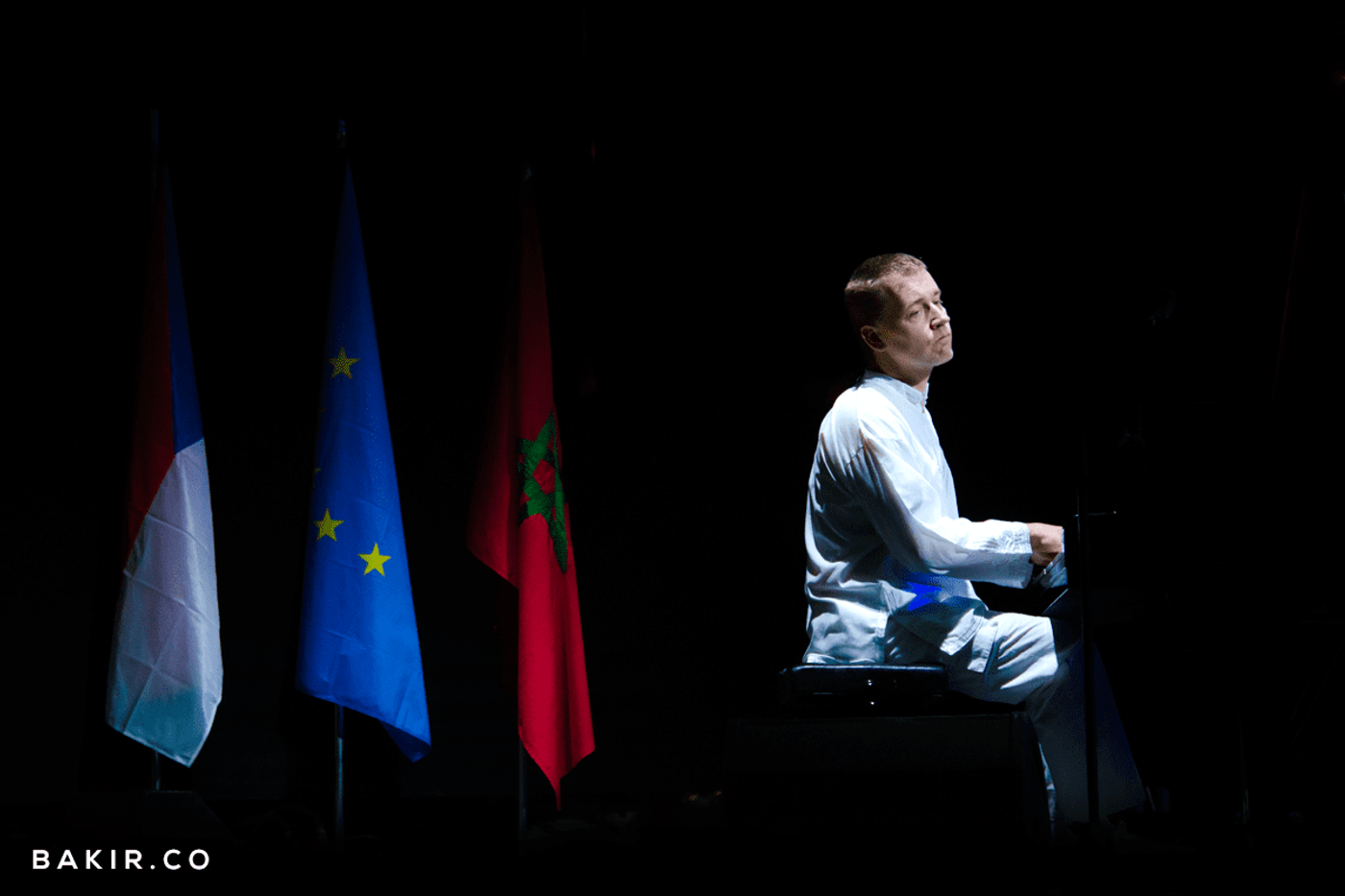 Pianist in white plays a grand piano on a dark stage beside Luxembourg, EU and Moroccan flags