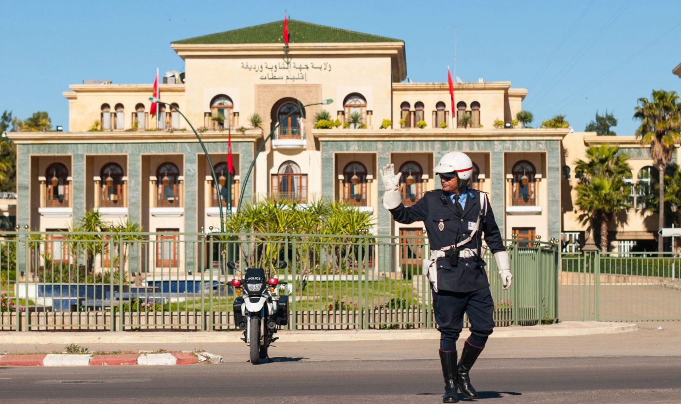 Moroccan traffic officer in white helmet directs traffic outside a provincial government building, police motorcycle parked behind