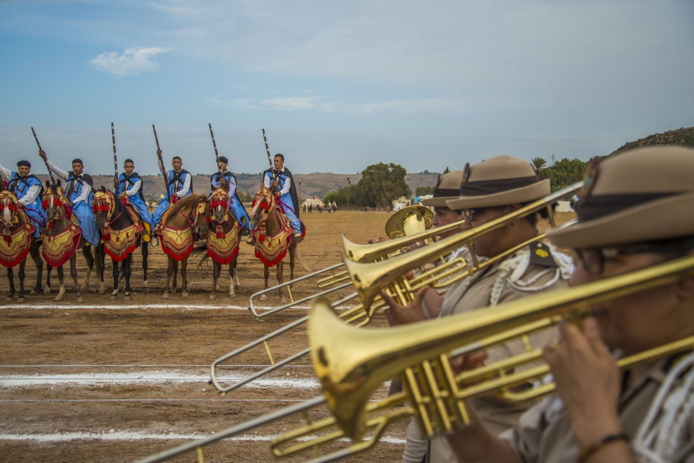 Horsemen in blue robes line up with raised rifles for a fantasia as brass band trombonists play in the foreground