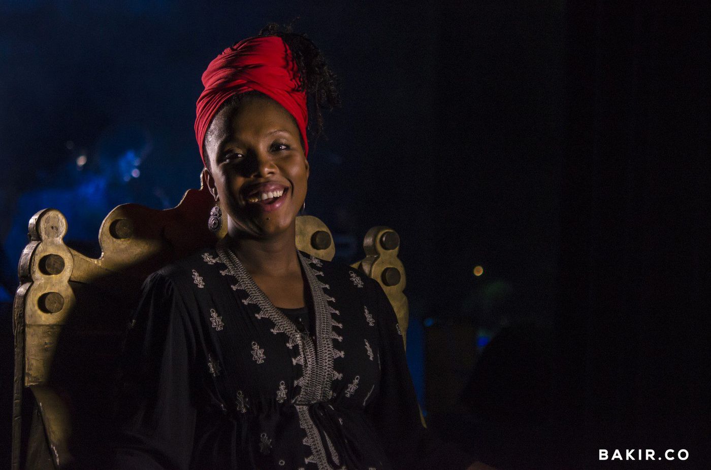 Smiling woman in a red headwrap and embroidered black dress seated on a carved gold chair