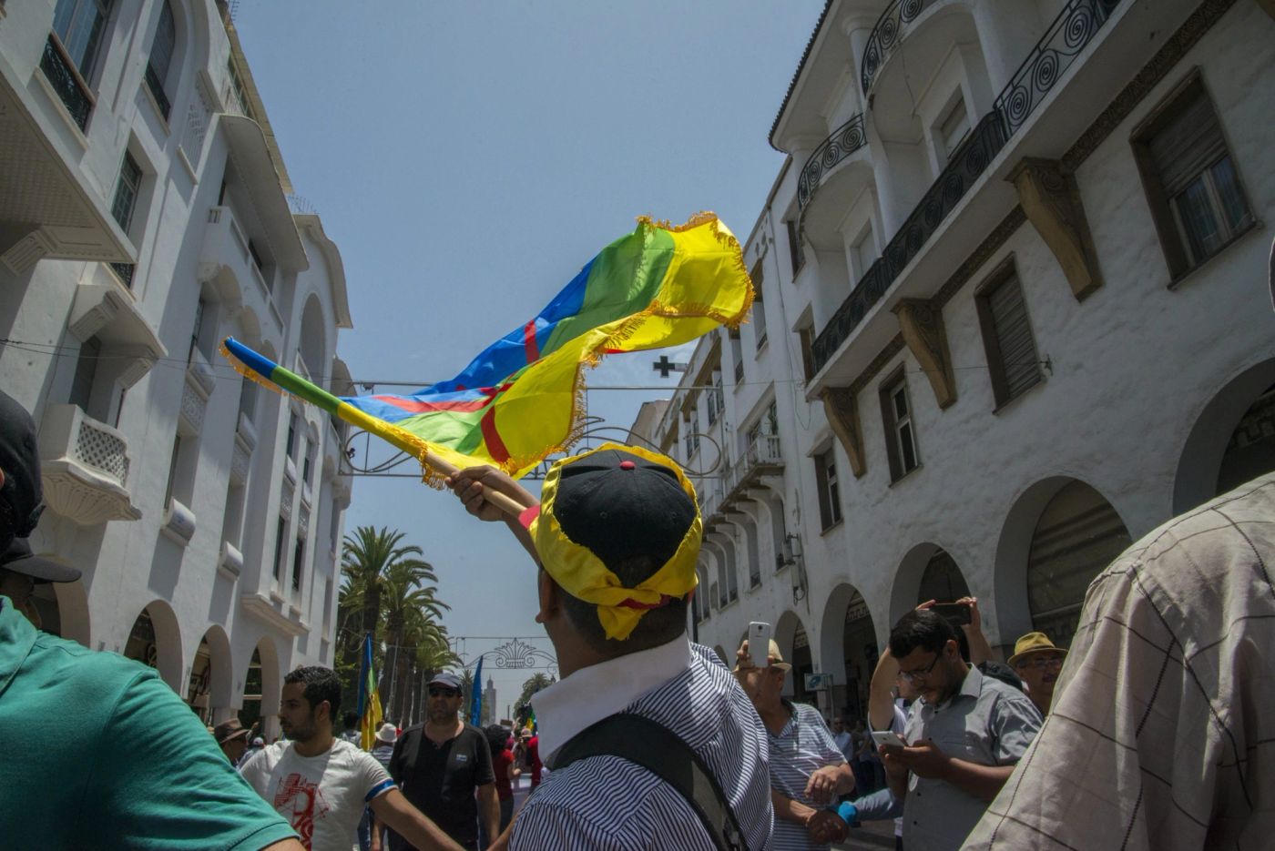 Demonstrator raises a large Amazigh flag amid a crowd marching between white buildings on a sunny street