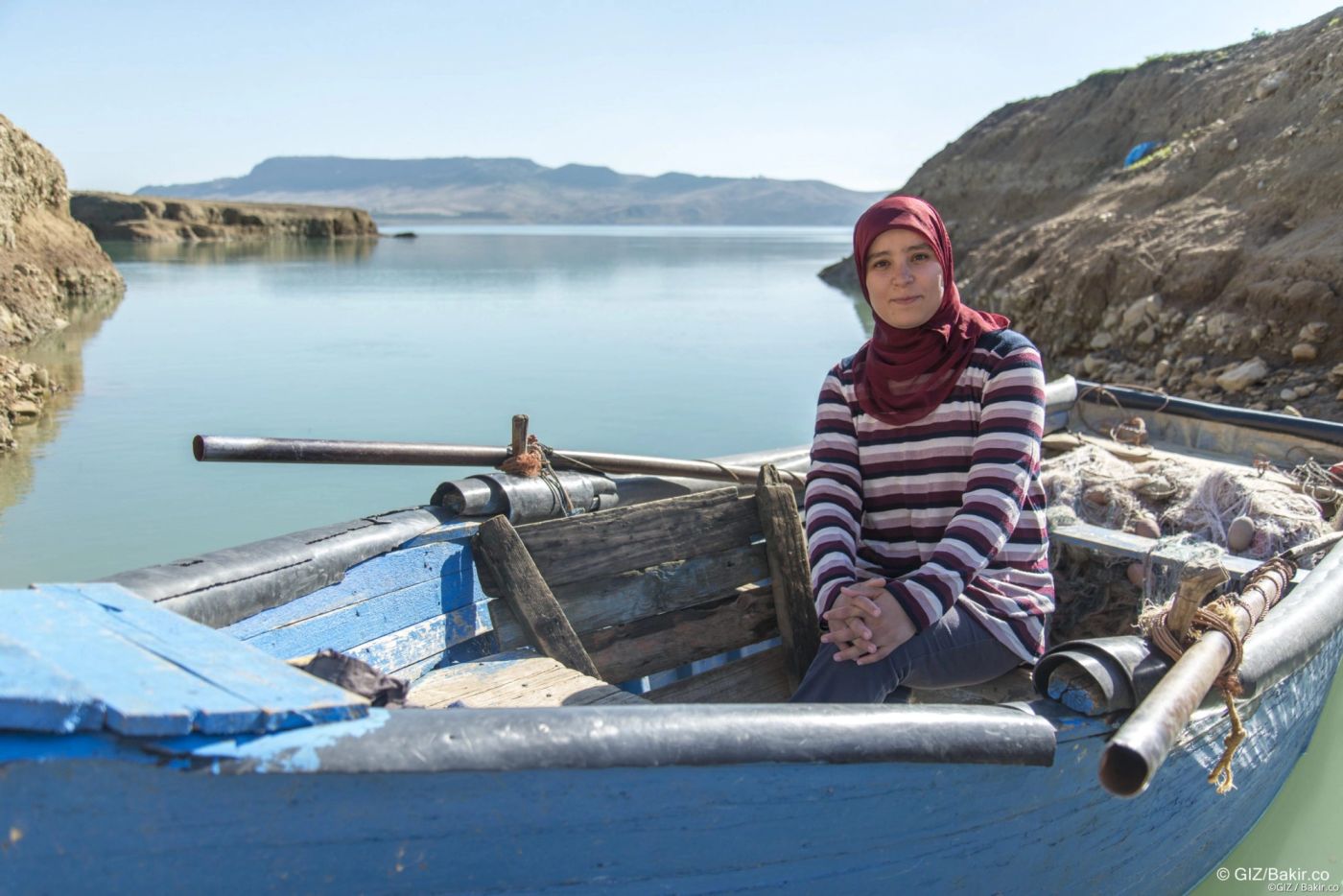 Young woman in red headscarf seated in a blue wooden fishing boat on a calm reservoir