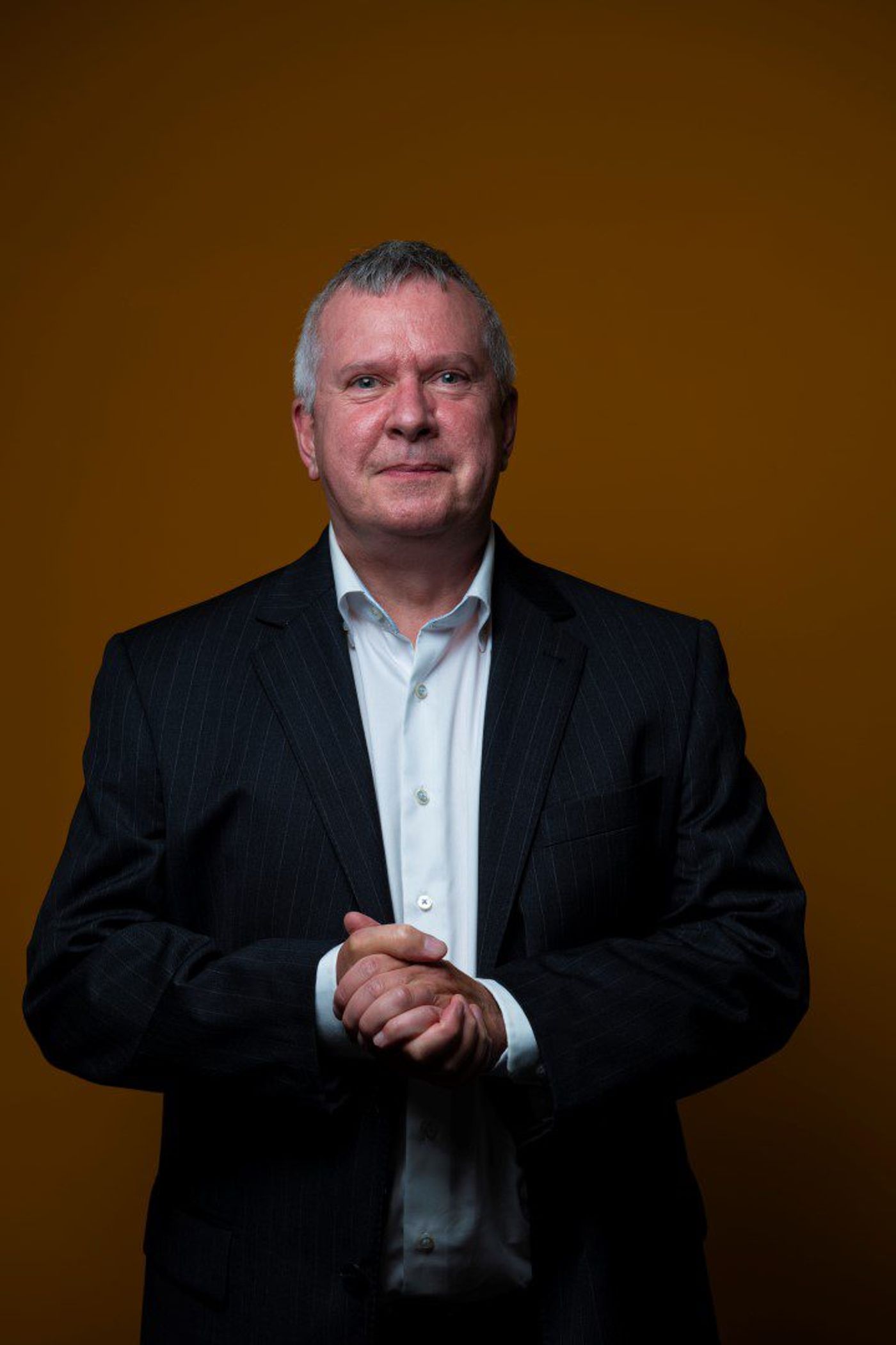 Grey-haired man in dark pinstripe suit, hands clasped, against an amber backdrop