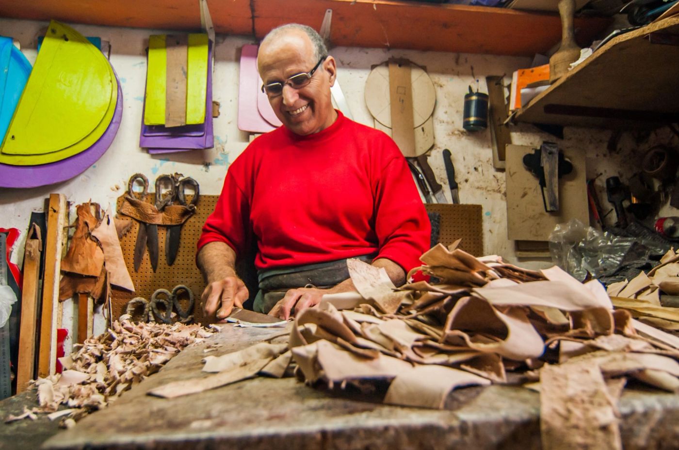 Smiling craftsman in red sweater cuts leather hides at a cluttered workshop bench