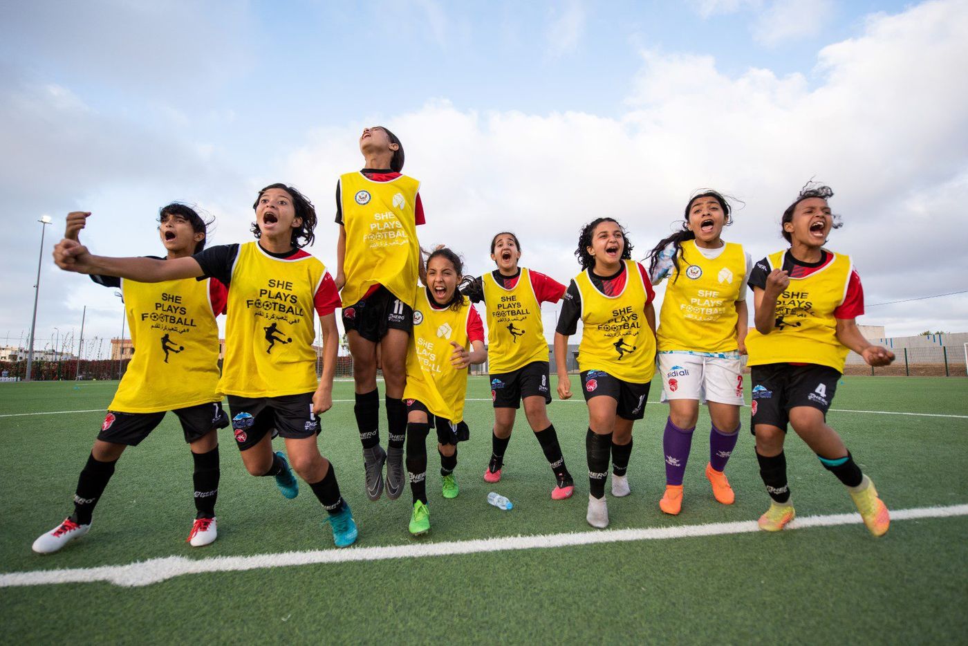 Girls in yellow She Plays Football bibs cheering and leaping on a turf pitch