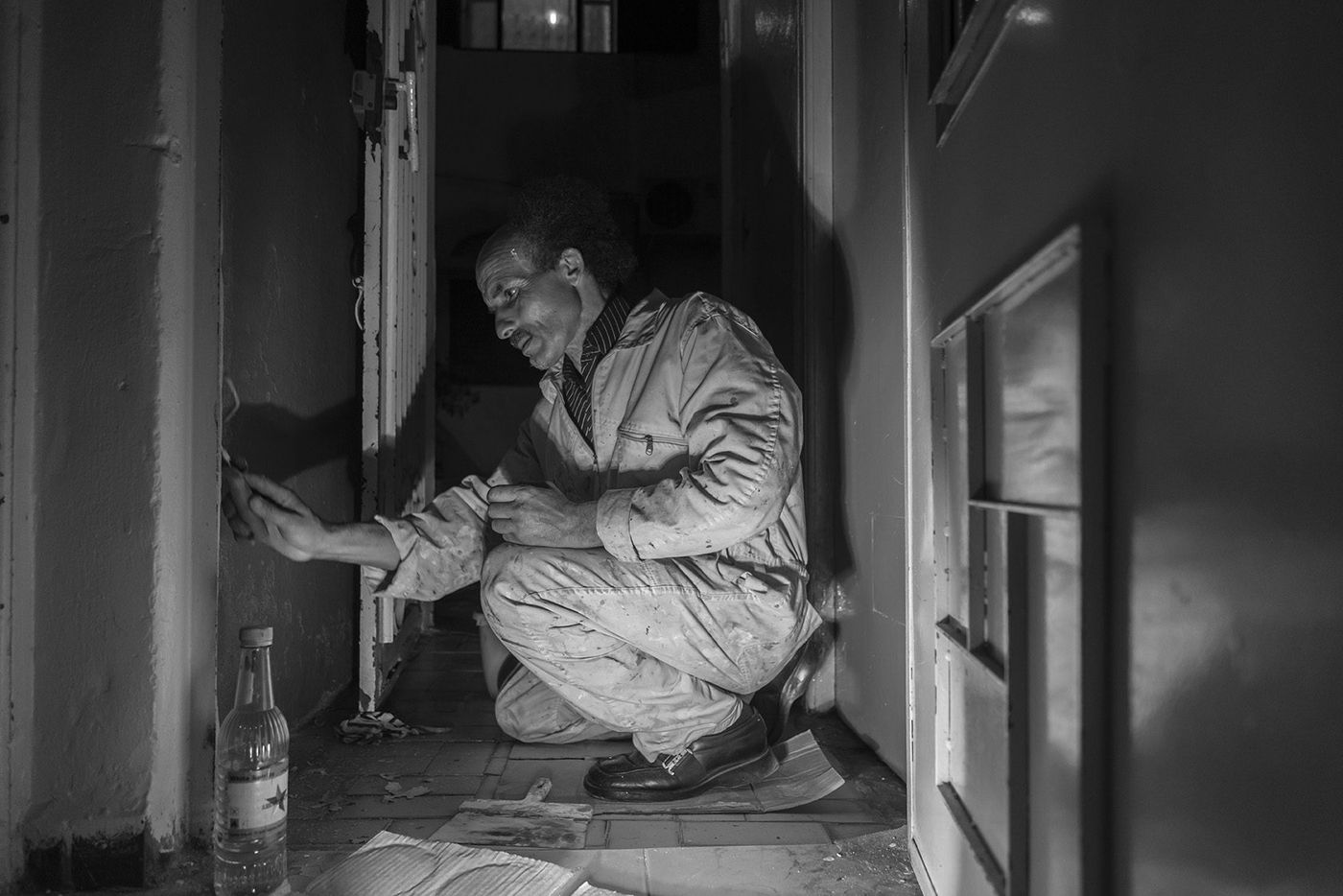 Man in paint-stained coveralls crouches in a dim doorway painting a wall, glass bottle on the floor, black and white