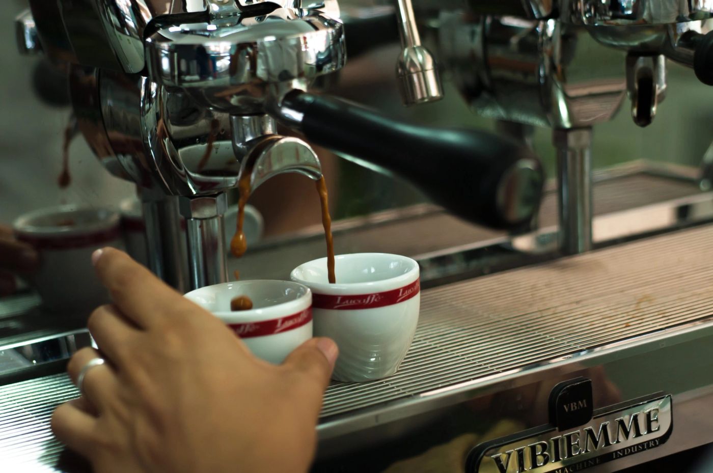 Espresso pours into two small branded cups under a chrome machine, hand steadying them