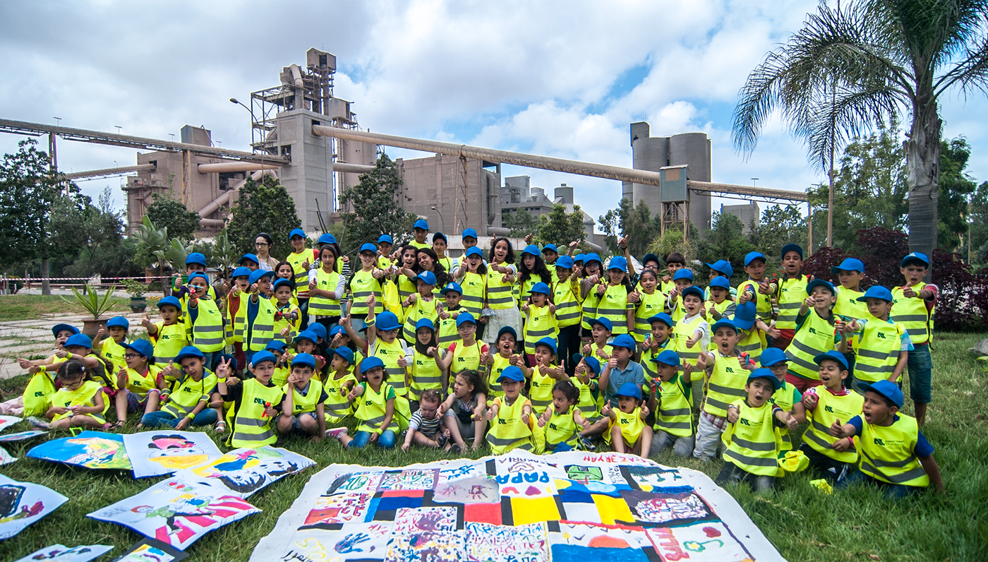 Children in yellow vests and blue caps pose with paintings before a cement plant
