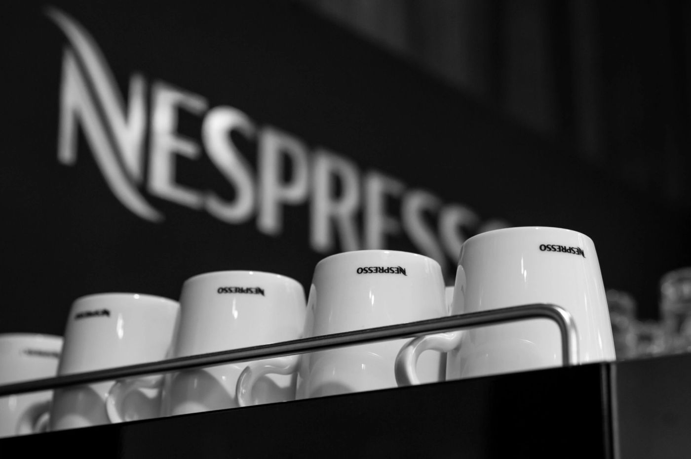 Rows of white Nespresso-branded cups stacked on a machine against a dark logo backdrop, black and white