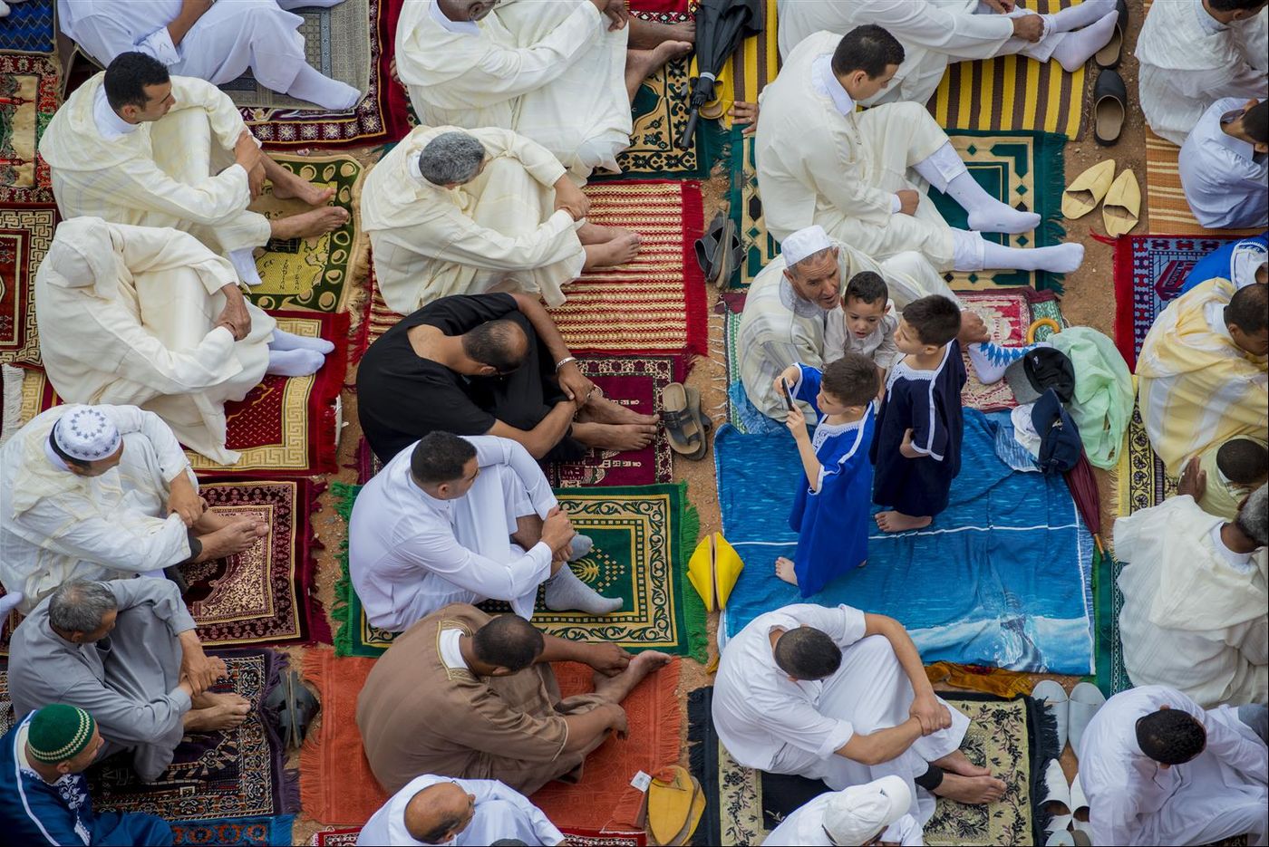Overhead view of men in white robes kneeling on colorful prayer rugs for Eid prayer, boys gathered to the side